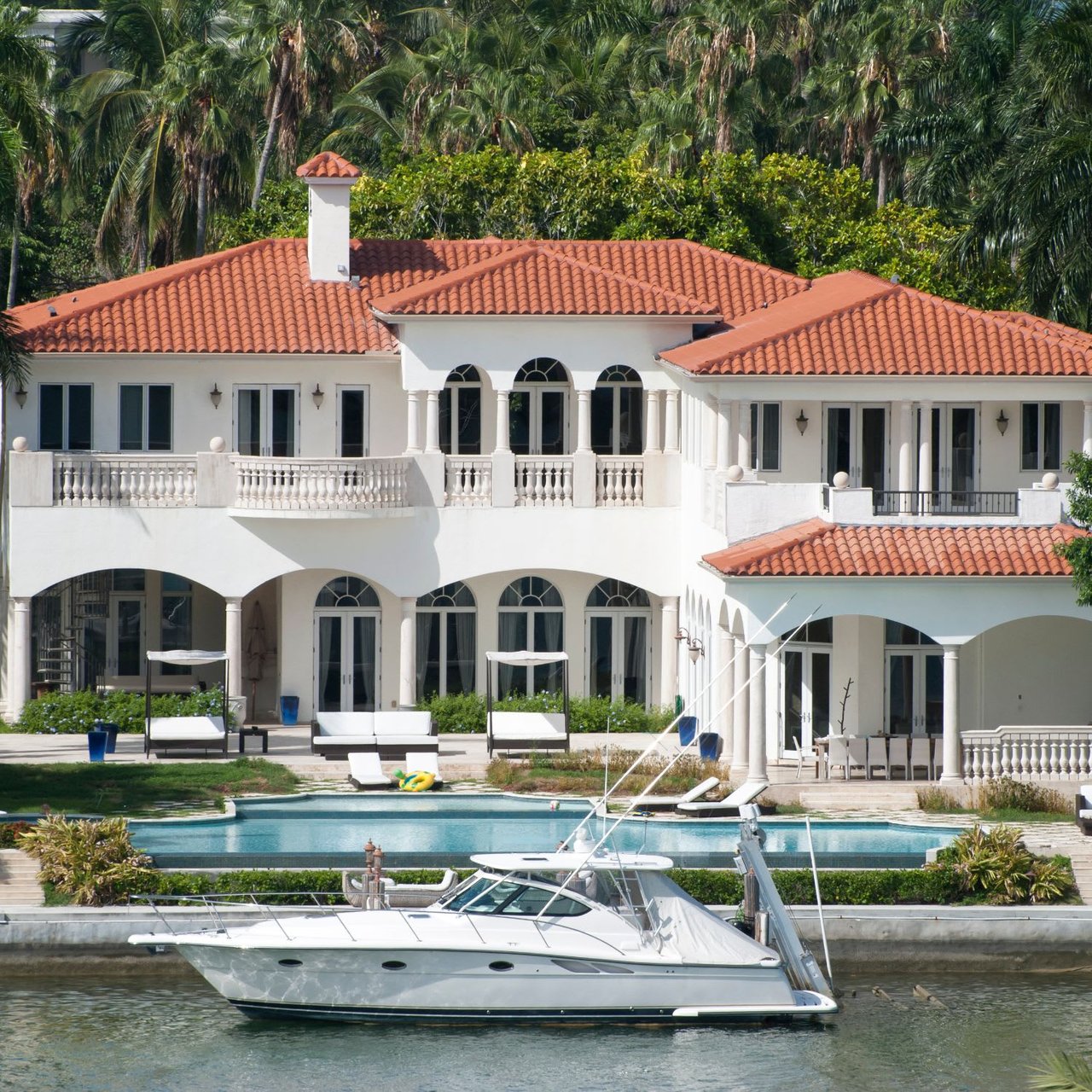  A grand white mansion with a red tile roof sits on the edge of a calm body of water.