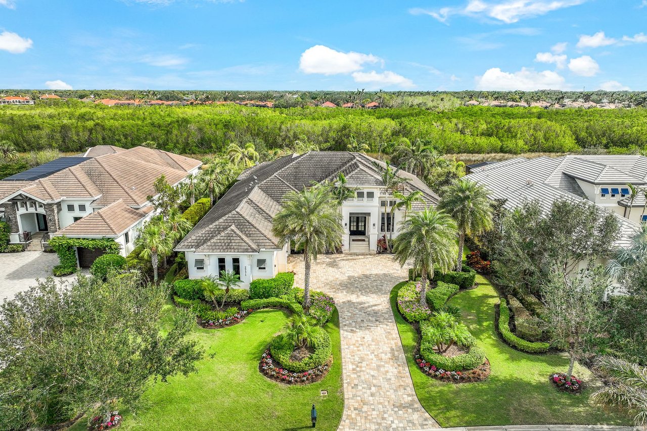  An aerial view of a large, luxurious white house with a dark tiled roof, situated amidst a lush green landscape.
