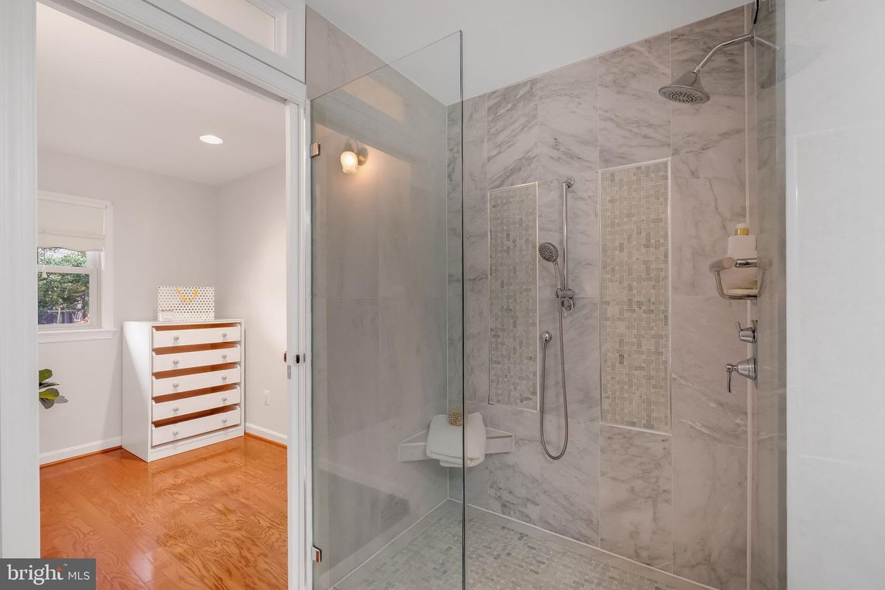 Marble-tiled walk-in shower with glass enclosure and mosaic accent panels beside a hardwood-floored dressing area.