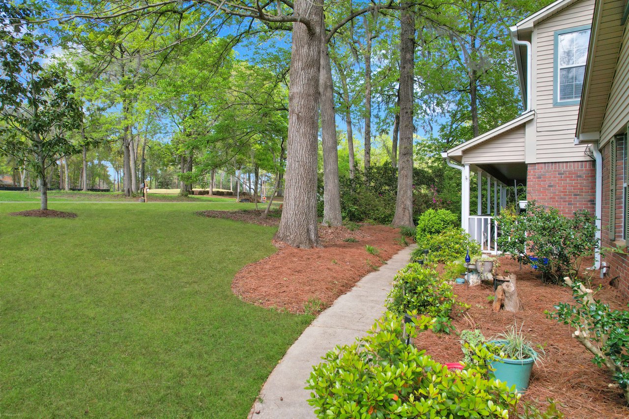 An exterior view of a house with a well-maintained garden and lawn. The house is surrounded by trees and has a picturesque setting.