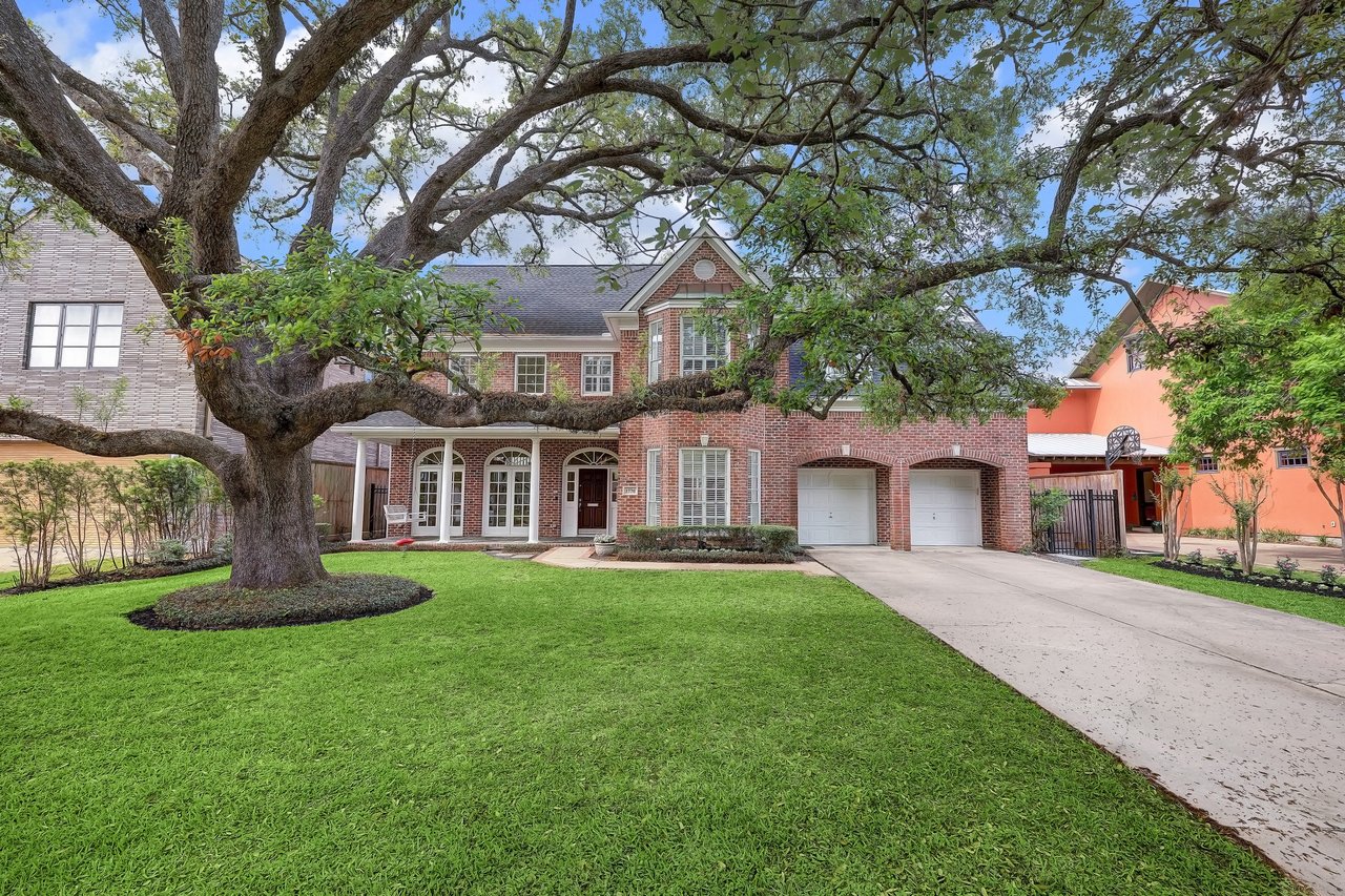 A two-story red brick house with a large front yard, a porch, a paved walkway, and a two-car garage in a quiet neighborhood.
