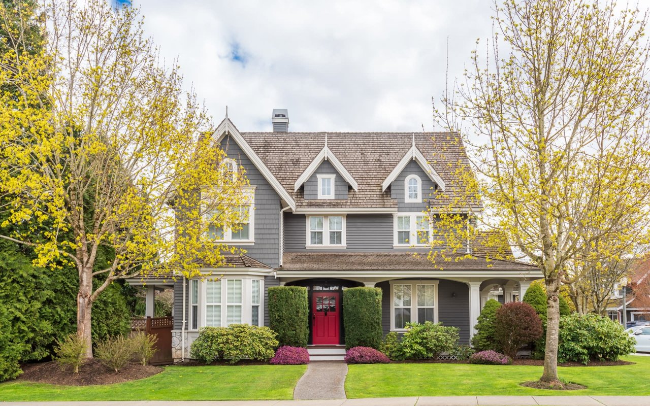 A large, two-story house with a red front door, walkway, covered porch, trees on the front lawn, and side garage.