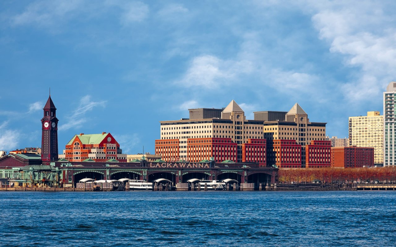 Hoboken, New Jersey's waterfront area with modern high-rises and older buildings, a clock tower, and a bridge over water.