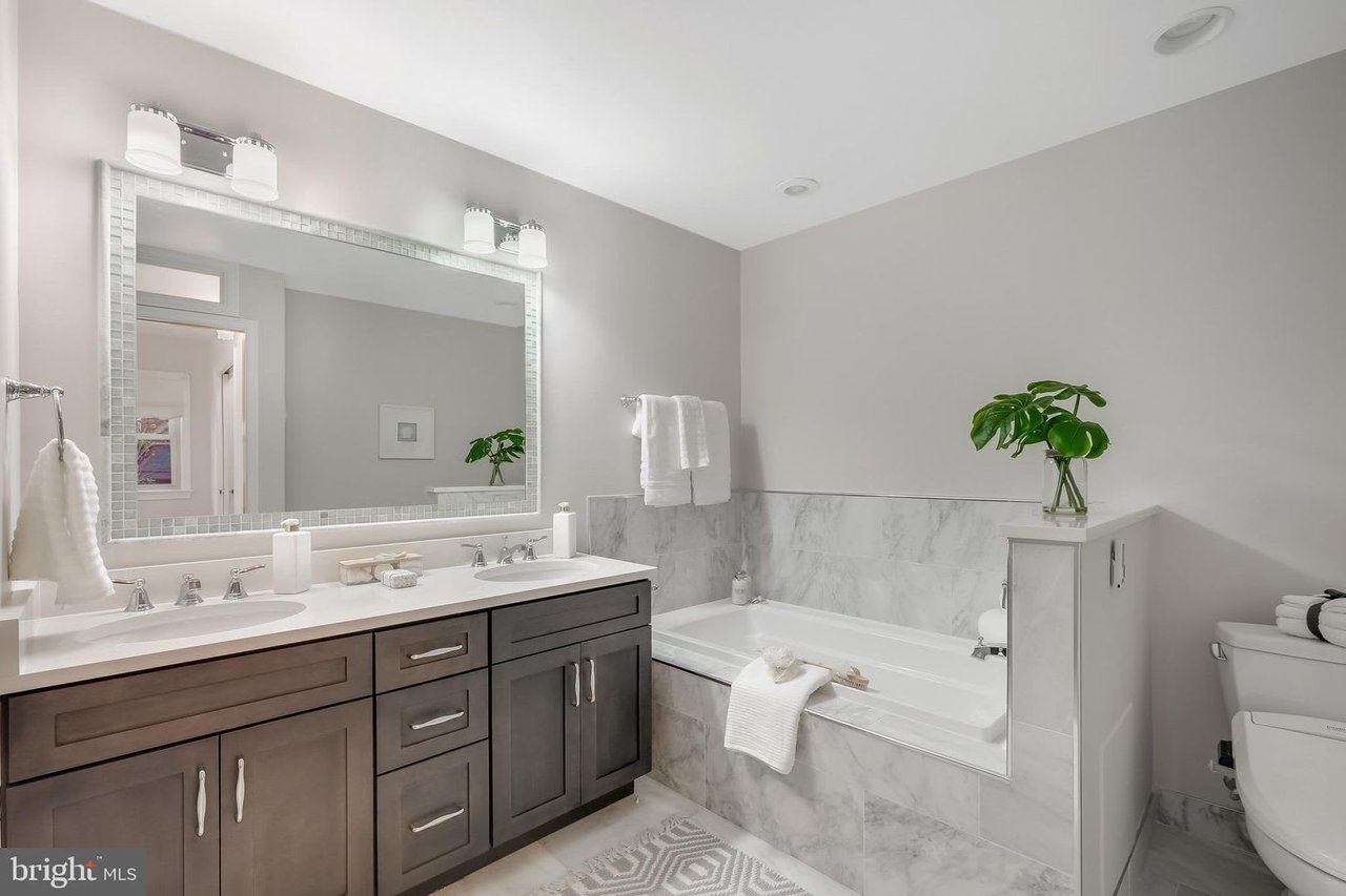 Bathroom with a dark double vanity, large mirror, and marble-look bathtub surround.