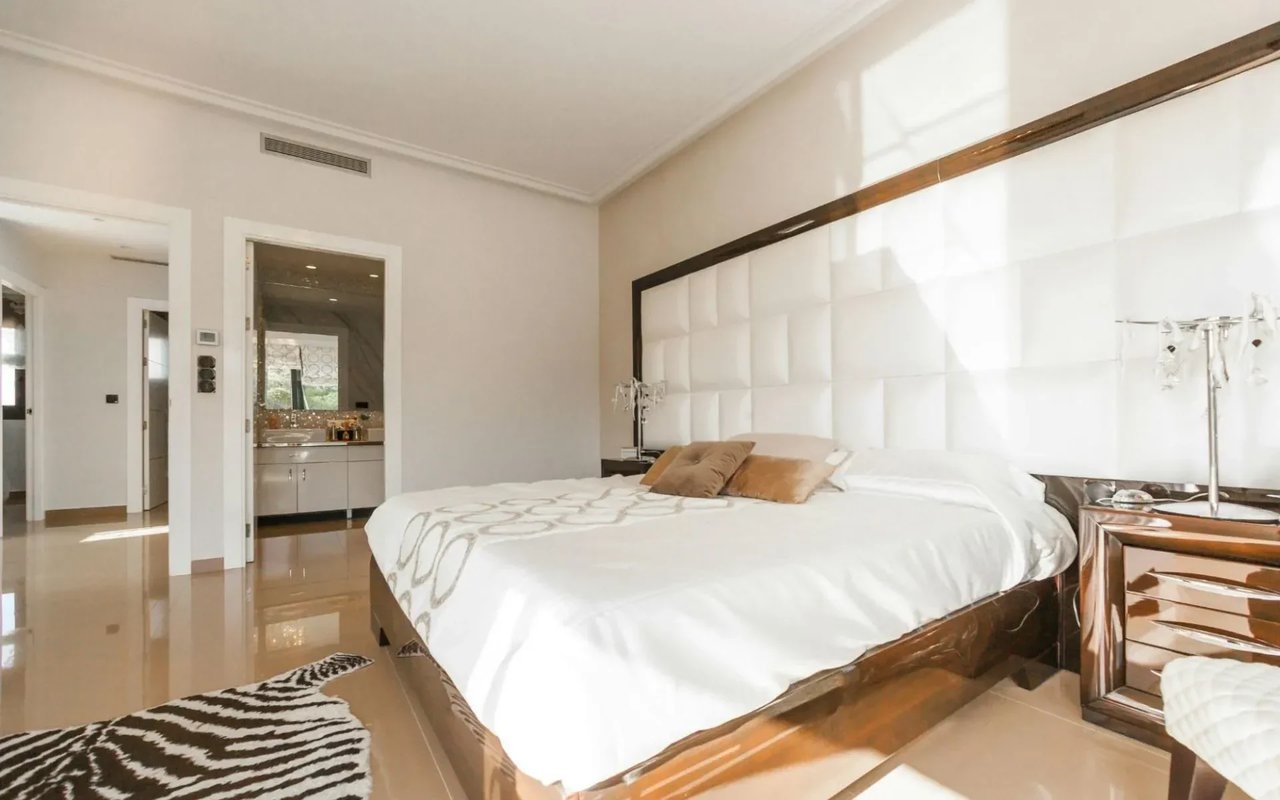 A modern bedroom with a white headboard, a bed with white linens, a zebra-print rug, and a visible glass-walled bathroom.