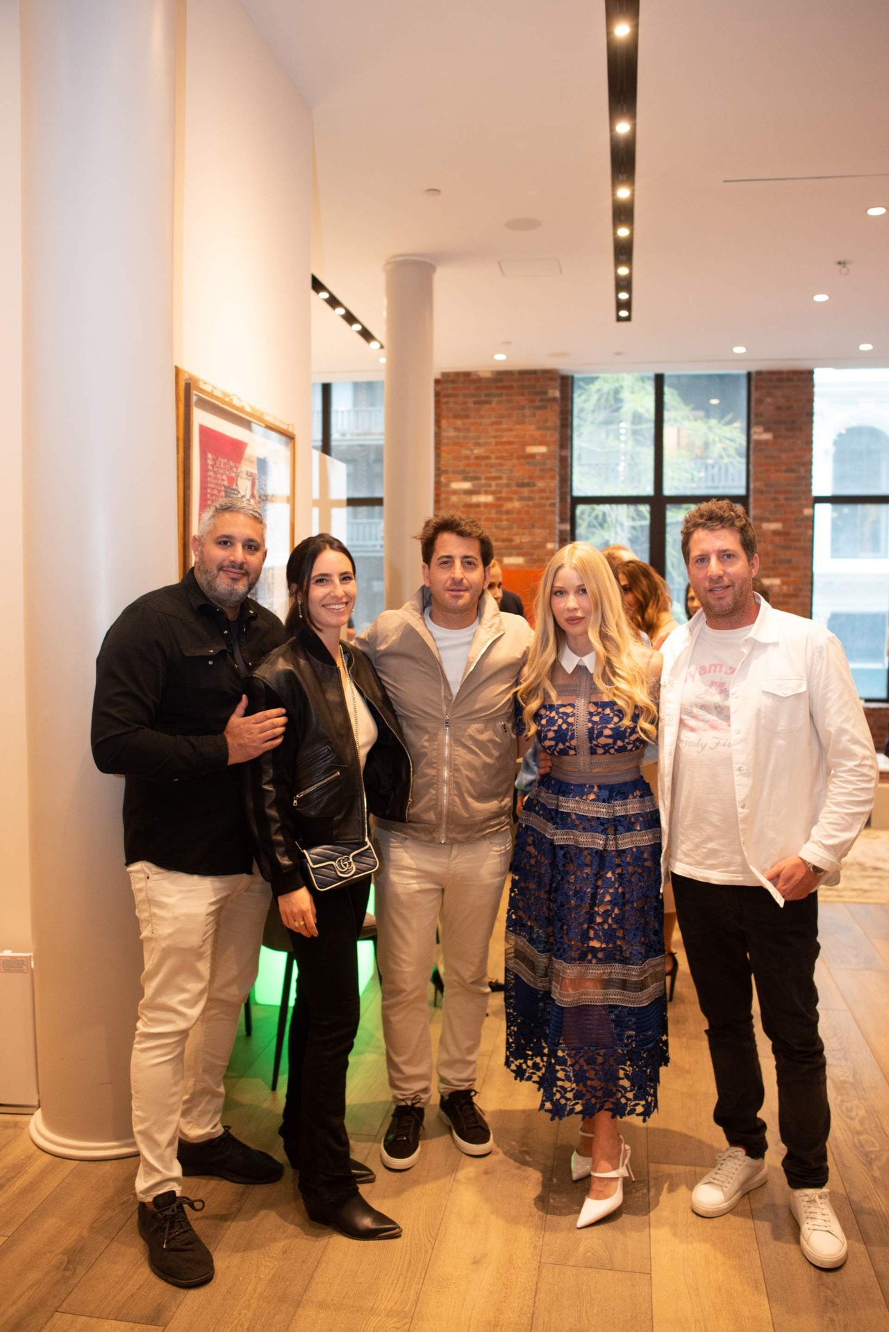 Five adults pose together in a bright event space, with brick walls and large windows behind them.