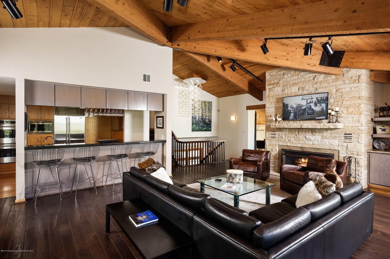 Open-plan living room and kitchen with vaulted wood ceiling, leather seating, and a stone fireplace.
