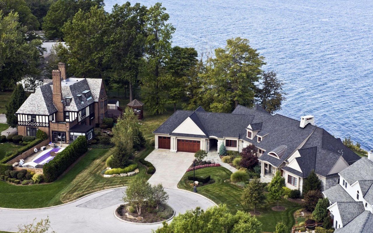 Aerial view of a large house with a swimming pool next to a lake. The house is white and has a red roof. The swimming pool is blue and has a white border. The lake is green and has a sandy beach.