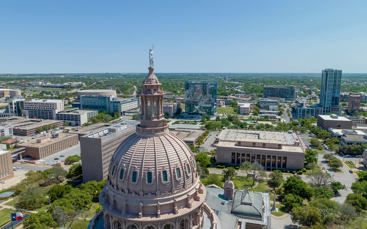 An aerial view of the Texas State Capitol in Austin with a large green space in front and an overlooking view of the city.