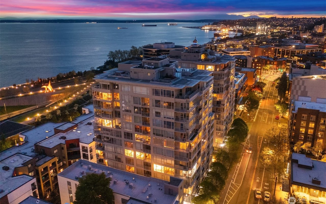 Aerial view of Seattle waterfront and condos illuminated at night, with a sunset sky in the background.