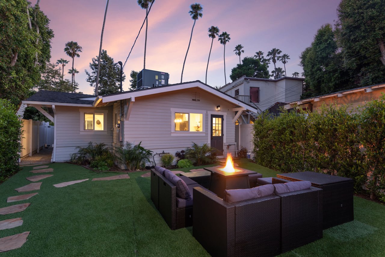 A charming, light-colored bungalow with a dark roof and a red brick foundation sits nestled among lush green trees and shrubs.