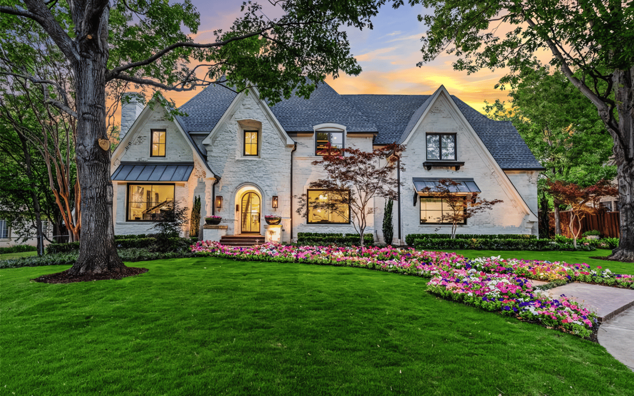 A  Colonial-style white house with a black slate roof, chimney, covered porch, walkway, front lawn, trees and flowers.