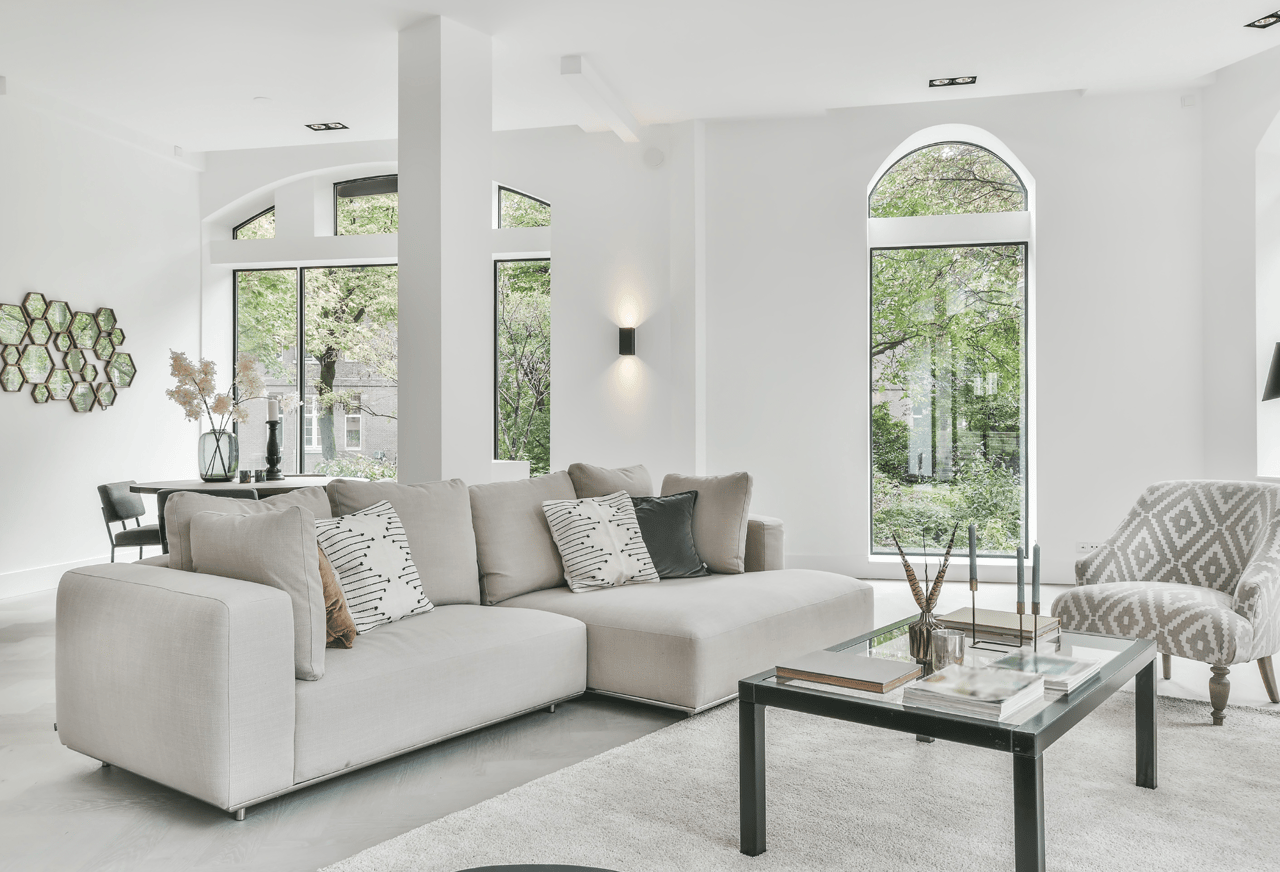 Bright living room with a gray sectional, patterned chair, glass coffee table, and tall arched windows.