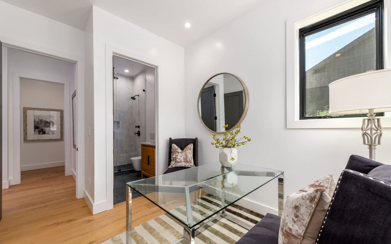 Living room with a glass coffee table, dark sofa, round mirror, and open doorway to a tiled bathroom.