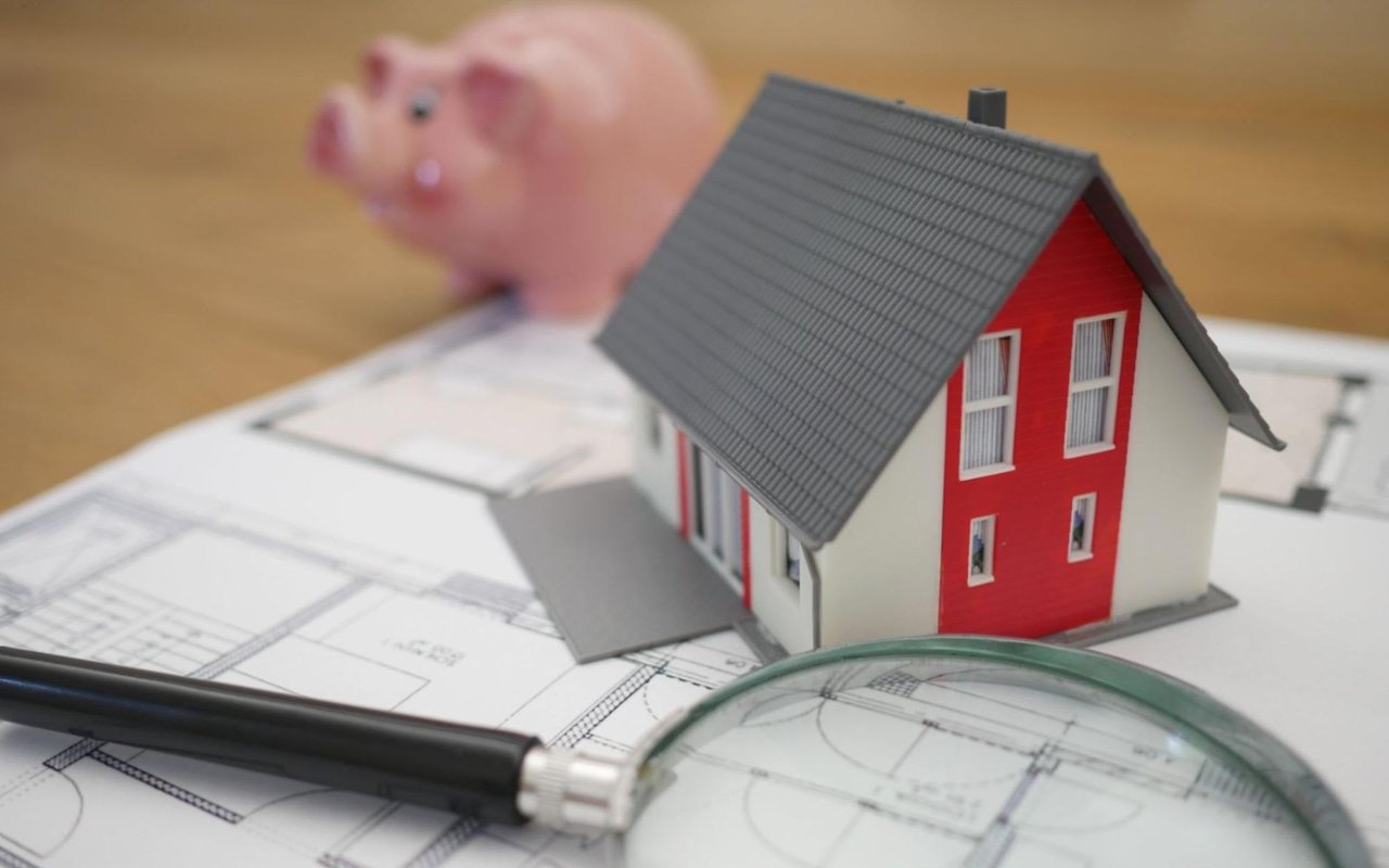 A small house model sits on a blueprint with a magnifying glass, a piggy bank in the background.