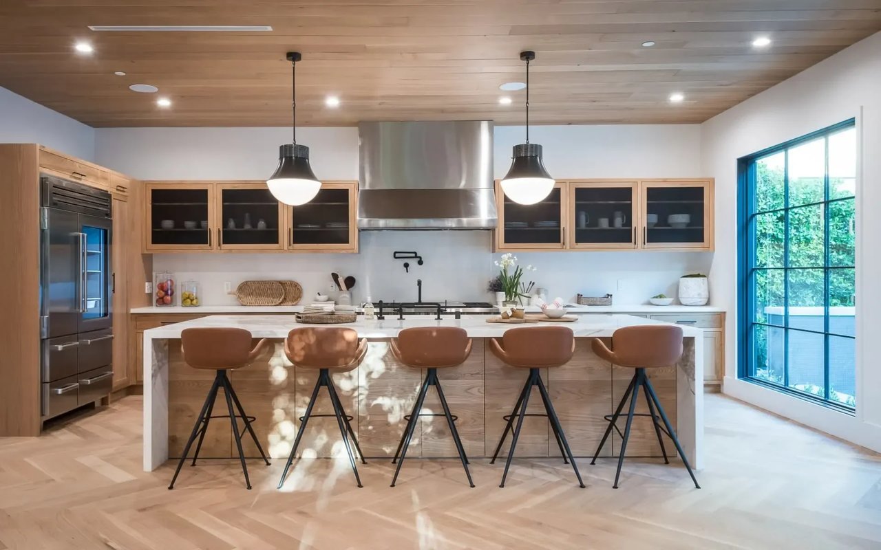 A modern kitchen with white cabinets, a large marble island with leather bar stools, and a wood-paneled ceiling.