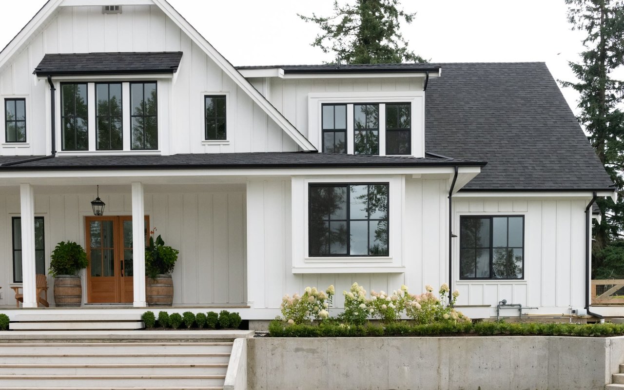 Raleigh homes for sale. A white house with a black roof, black dormer windows, a porch, and a picket fence surrounded by various plants and trees.