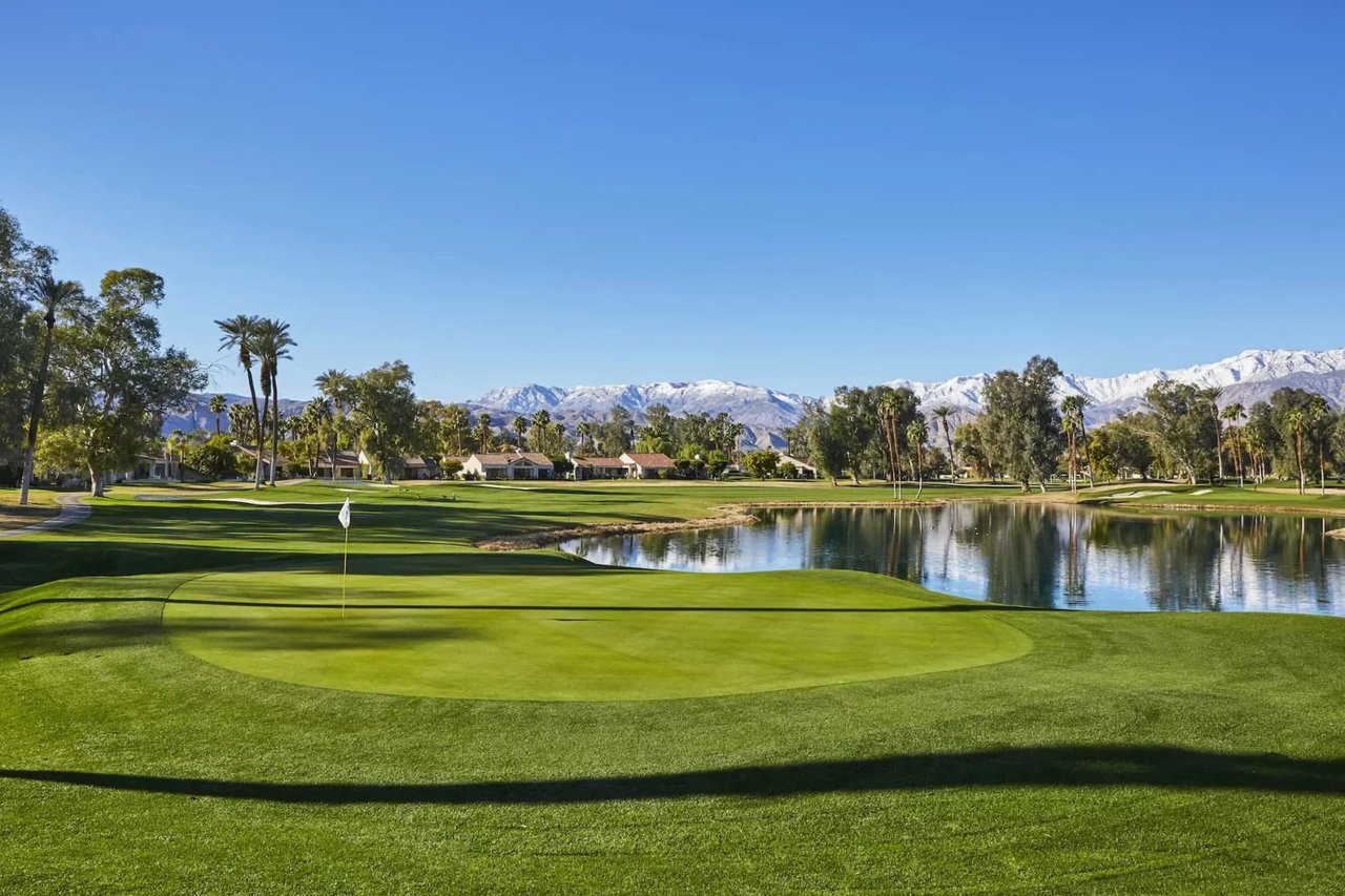 A golf course green with a flag and a pond in the foreground with mountains in the background