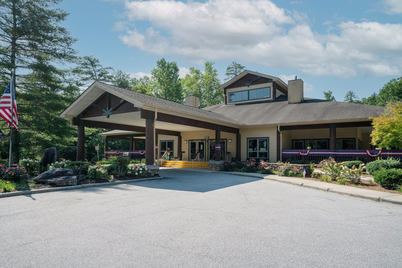 alt=A single-story building with a covered entrance, surrounded by landscaped gardens and an American flag in the foreground, serves as a vibrant platform offering career resources for real estate agents under a partly cloudy sky.