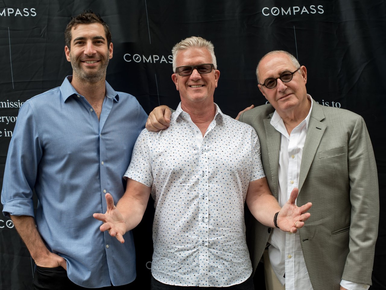 Three men are posing for a photo in front of a dark backdrop with the word "COMPASS" repeated.