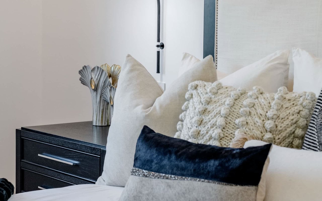 Staged bedroom with layered cream and navy pillows and a black bedside table with a sculptural vase.