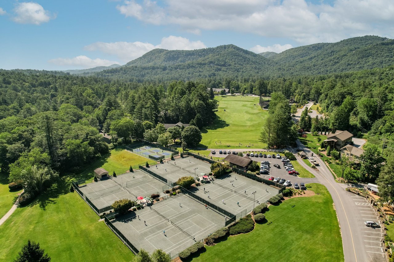 Aerial view of a sports complex featuring several tennis courts, a golf course, and parking area, surrounded by lush greenery and mountainous landscape in the background—an ideal visual asset for real estate agents showcasing premier properties.