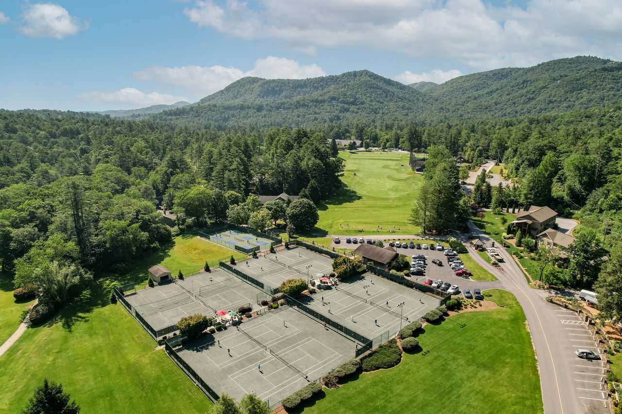 ALt=Aerial view of a sports complex featuring several tennis courts, a golf course, and parking area, surrounded by lush greenery and mountainous landscape in the background—an ideal visual asset for real estate agents showcasing premier properties.