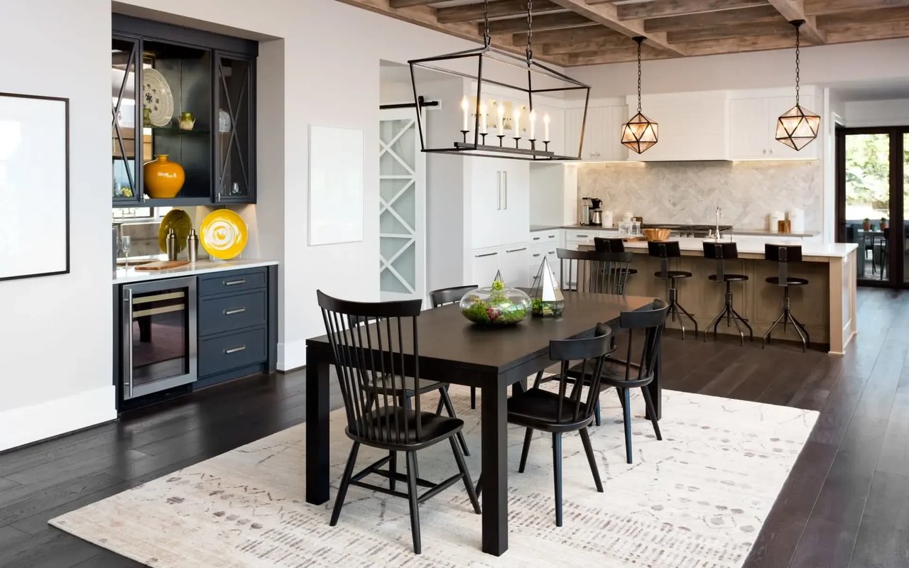 A  kitchen and dining room with white cabinets, a dining table, a black chandelier, a large island,  and a wine cooler.