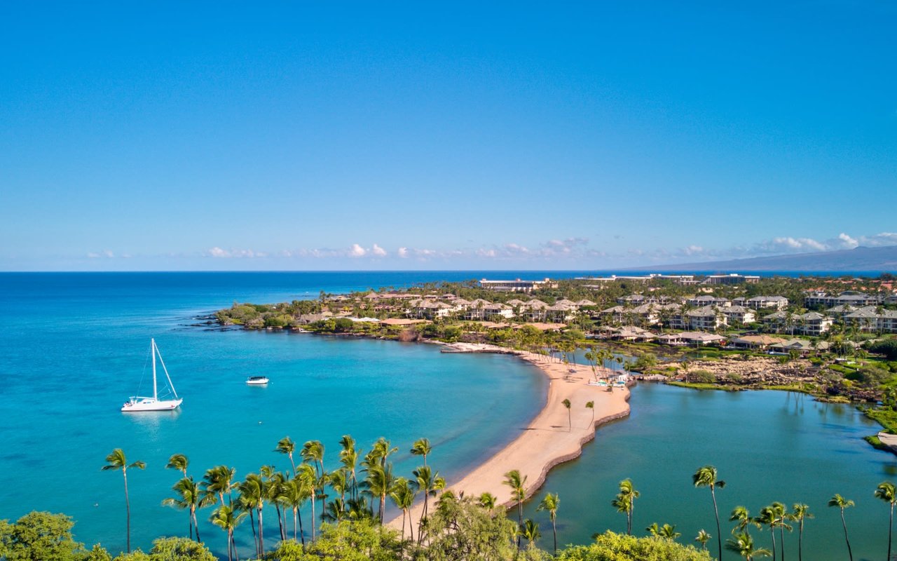 An aerial view of a sandy beach with a white catamaran sailboat in the clear blue water