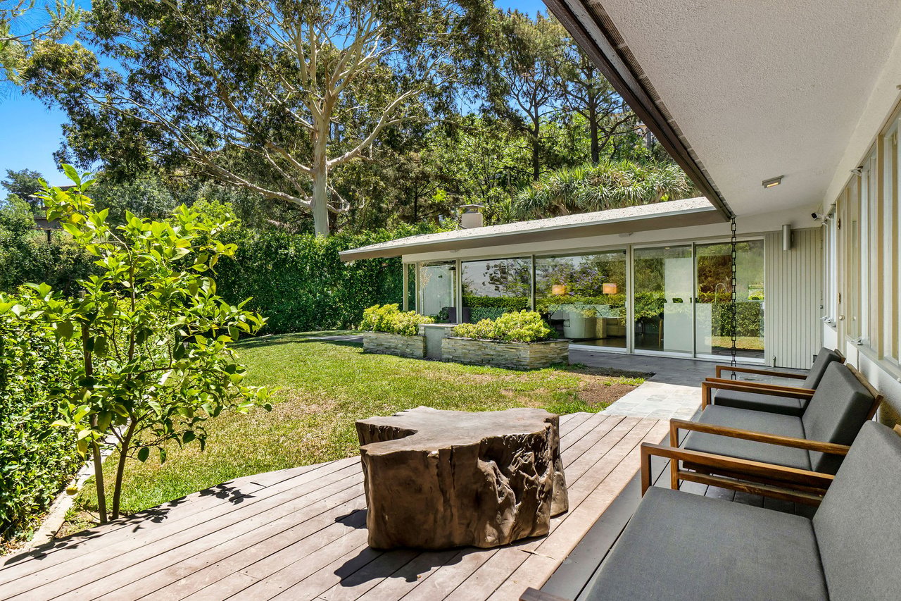 Backyard patio with a wood deck, cushioned chairs, and a tree-stump table beside large glass doors.