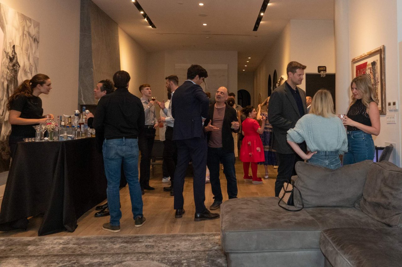 Guests mingle at a real estate gathering in a modern living room with a drinks table and gray sectional sofa.