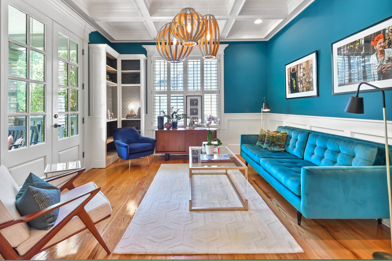 A blue-themed living room with a plush sectional couch, glass coffee table, blue upholstered chairs, and landscape paintings.