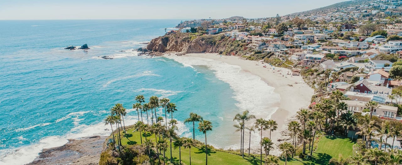 White sandy Laguna Beach with calm waves, tall and lush palm trees, and close to high-rise buildings.