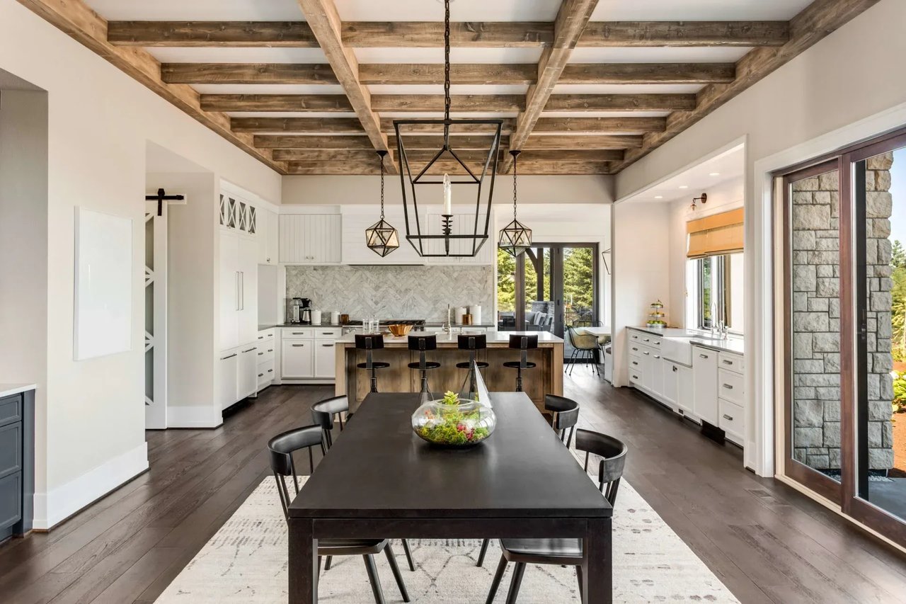 A modern kitchen with white cabinets, a dark wood dining table, a large island, and large windows overlooking a patio.