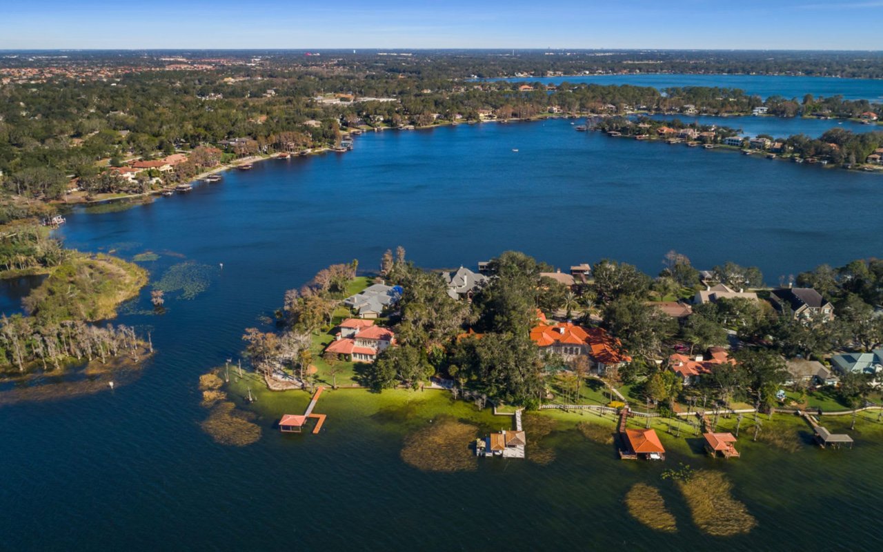 An aerial view of a lake surrounded by trees with colorful houses and docks built on its shores, along with some green areas.