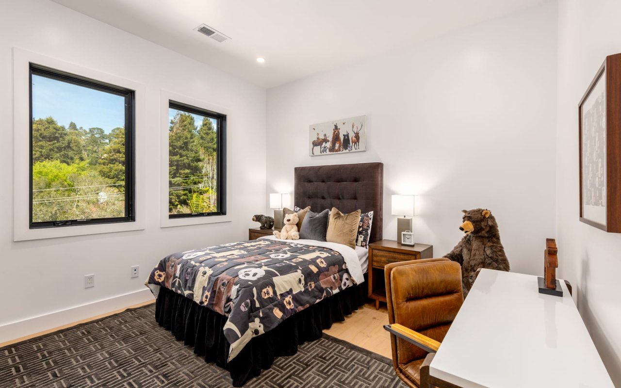 Bedroom with bear-print bedding, a tufted headboard, and windows overlooking trees.