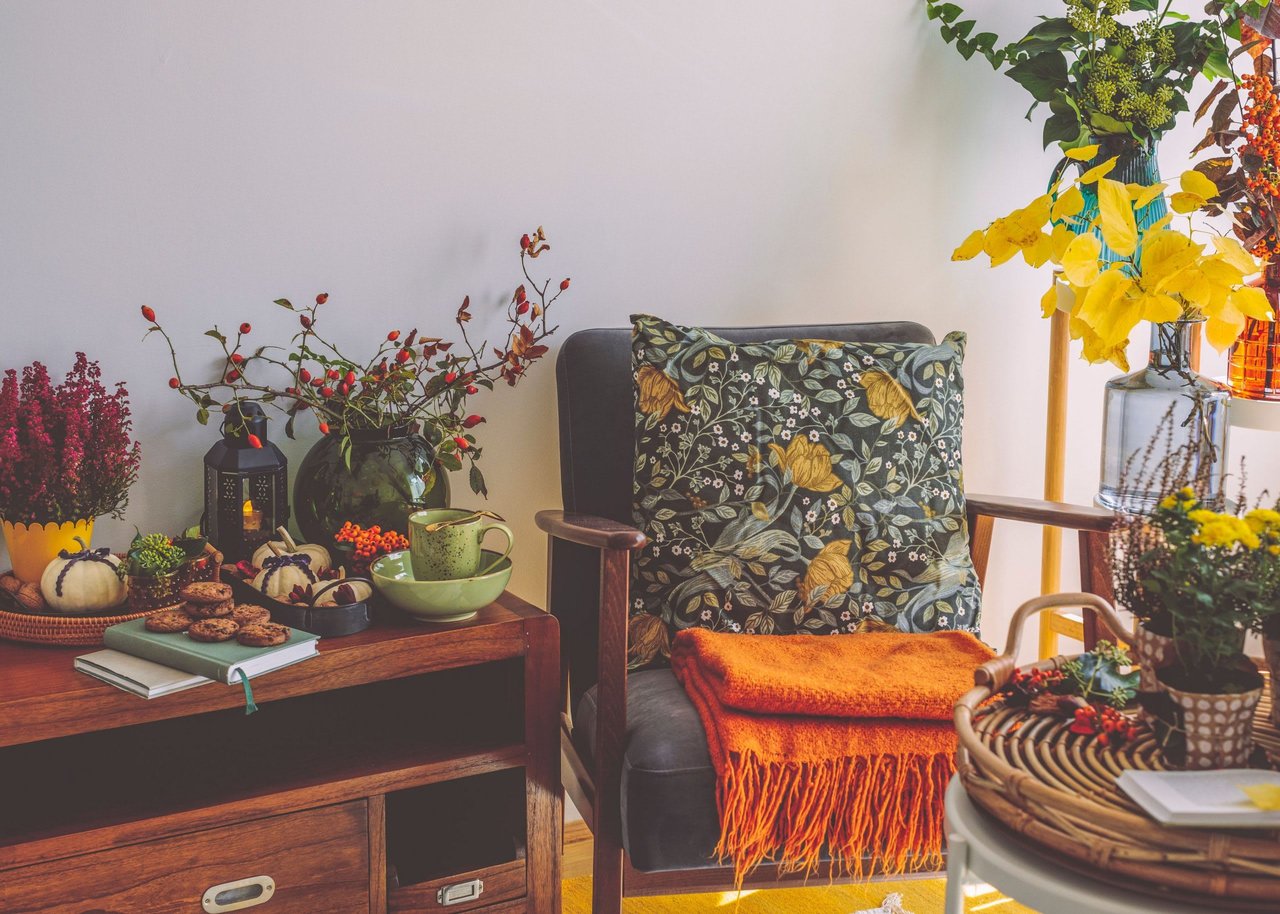 Fall seating nook with a floral armchair, orange throw, pumpkins, seasonal flowers, tea, and cookies.