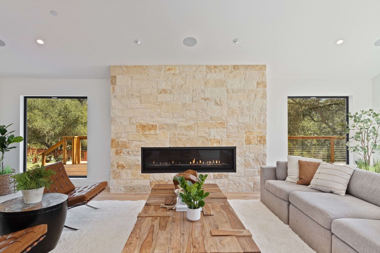 Living room with a stone fireplace, gray sofa, leather lounge chairs, and windows opening to a deck.