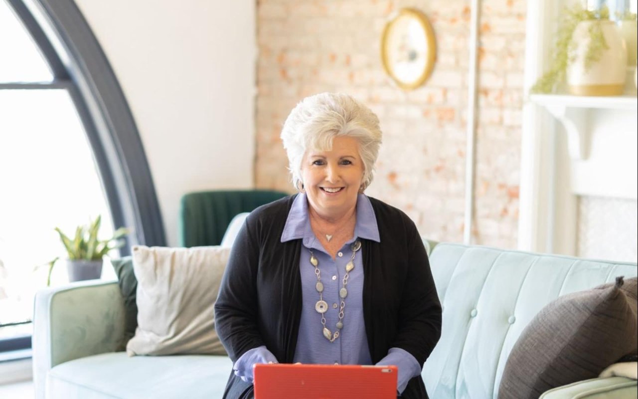 Smiling woman with short white hair sits on a pale blue sofa holding a red tablet.