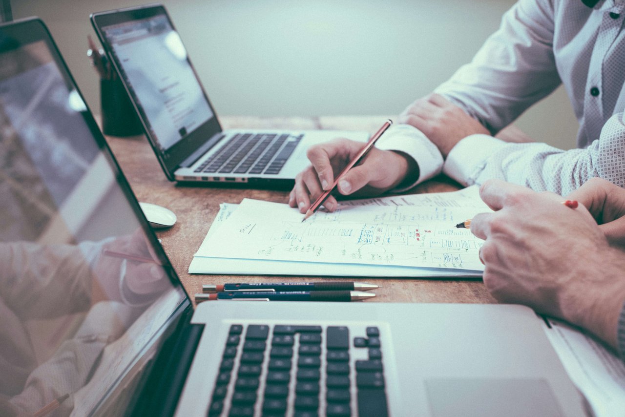 Two people sitting at desk flanked with computers referring to business document between them..