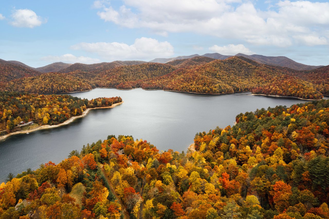 Aerial view of a lake surrounded by autumn-colored forests and mountains under a partly cloudy sky, an inviting landscape where real estate agents could network amidst nature's stunning backdrop.