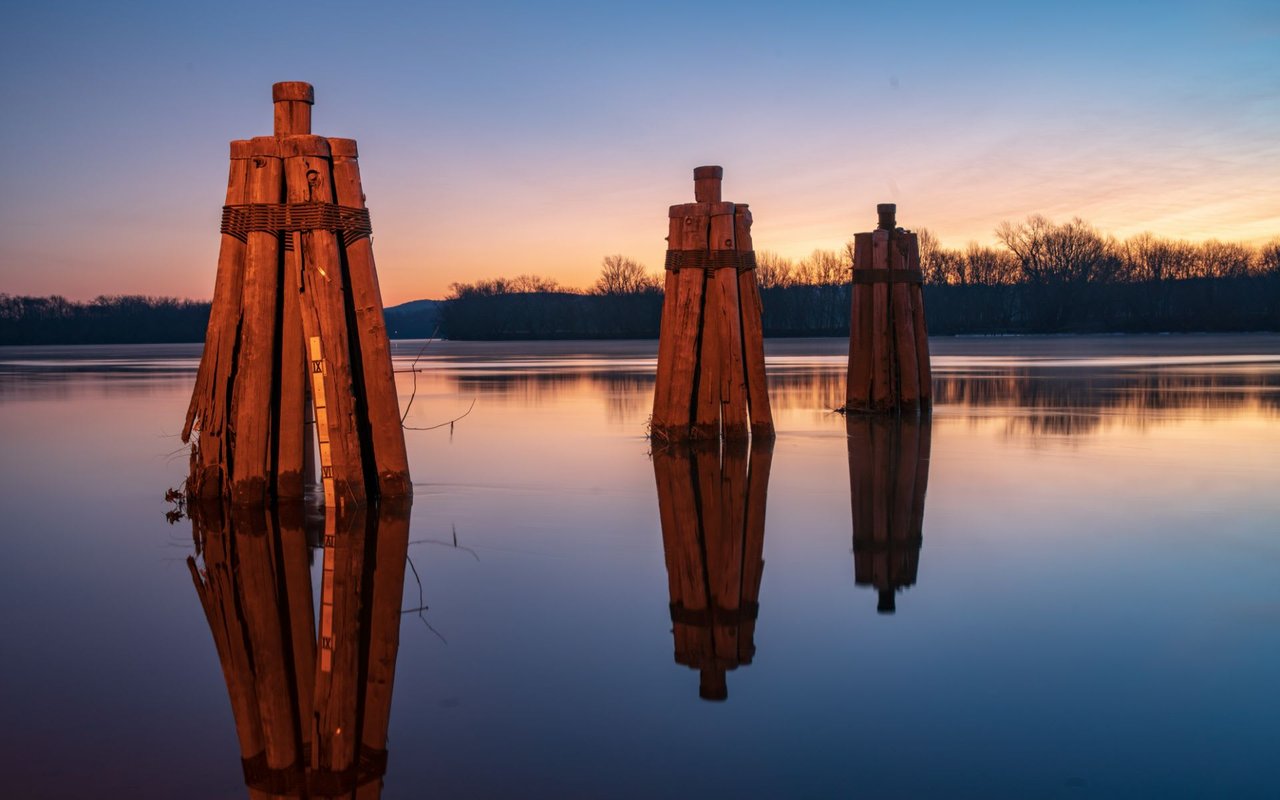 Sunset over the Connecticut River in Rocky Hill, CT, with three wooden posts partially submerged in the calm water, reflecting the warm colors of the sky and creating a serene and peaceful scene.