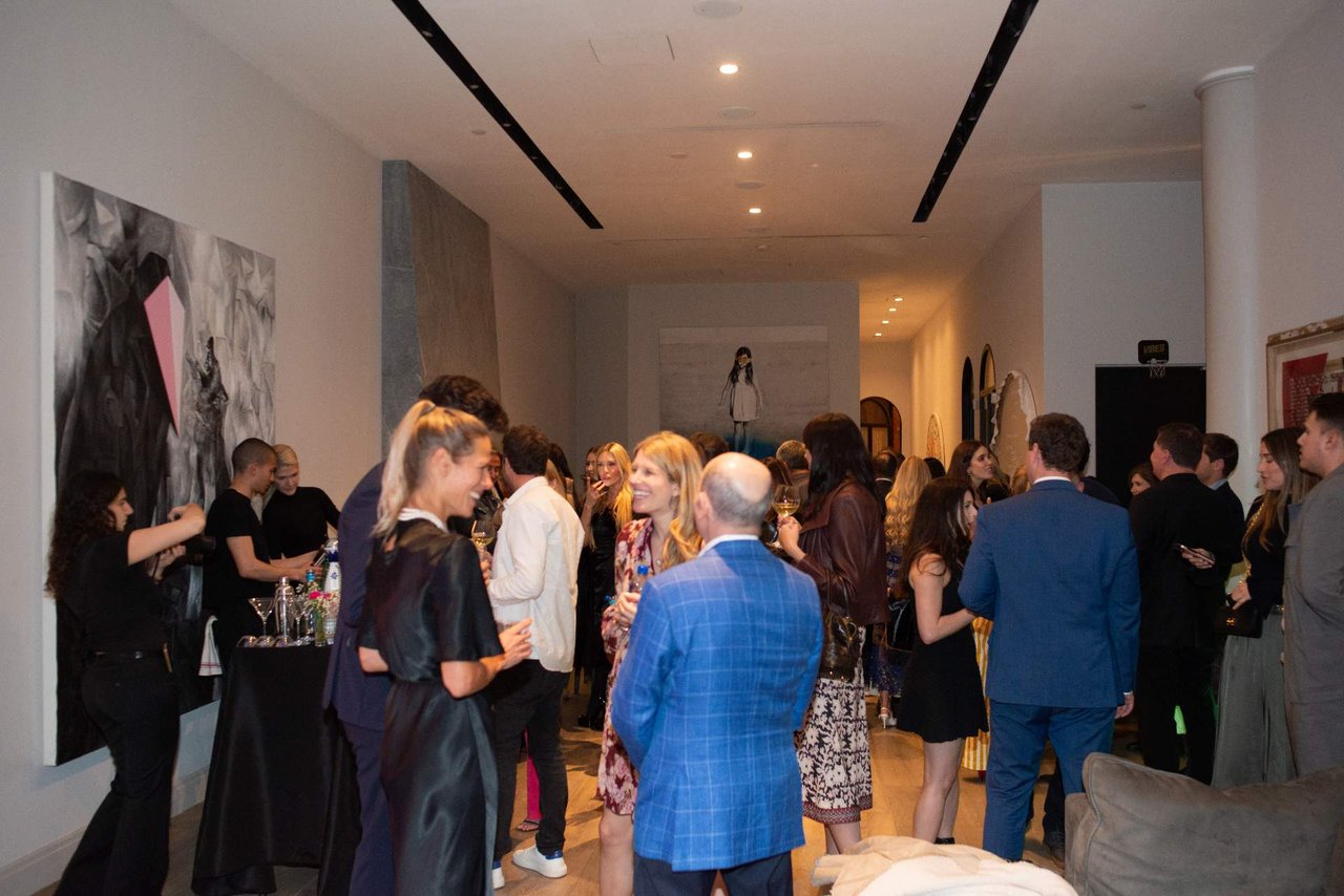Guests mingle at a real estate event beside abstract artwork and a drinks table.