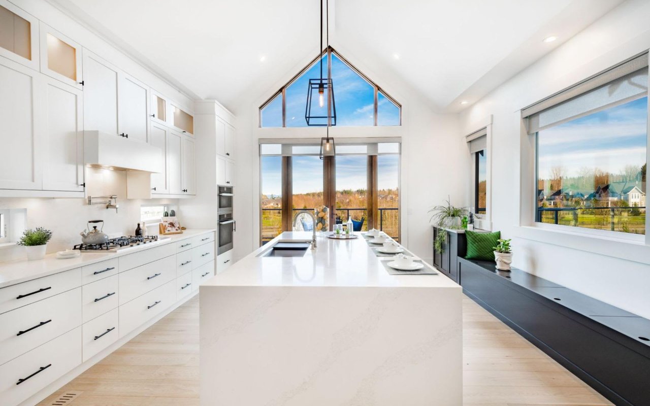 A kitchen with a large central island, light-colored countertops, stainless steel appliances, and outside-view windows.