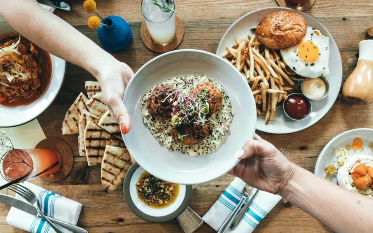  A high-angle shot captures a variety of dishes on a wooden table, with two pairs of hands in the foreground, one offering and one receiving a white bowl.