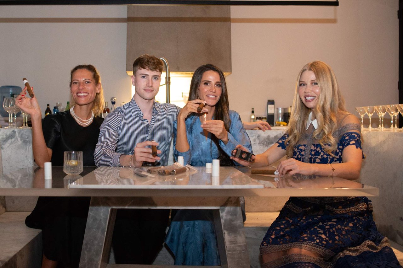 Four people pose together at a marble table, holding cigars at an indoor gathering.