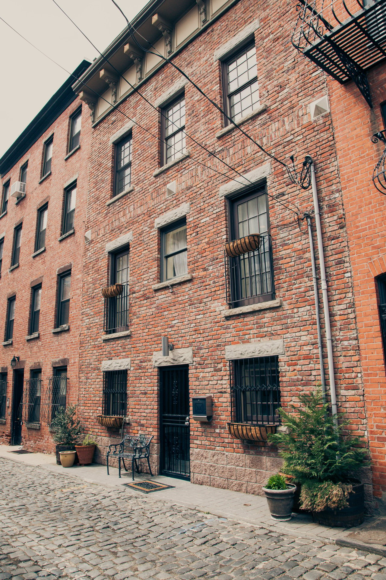 A large brick building on a cobblestone street with a flat roof, black metal fire escape stairs, and a front sidewalk.