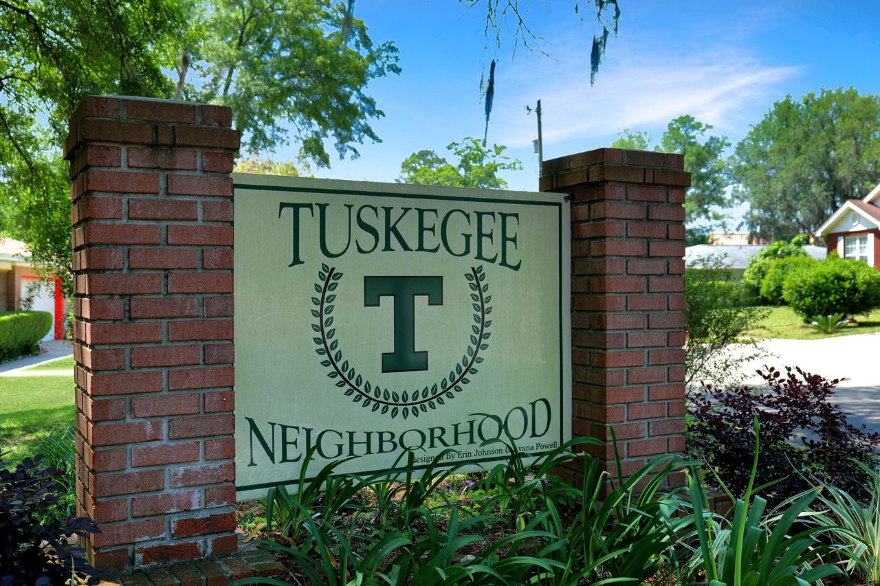 A close-up shot of a sign that reads "Tuskegee Neighborhood." The sign is set in a brick structure and surrounded by plants.