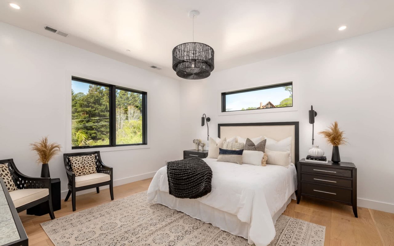 Bright bedroom with a white bed, black furnishings, accent chairs, and windows overlooking trees.