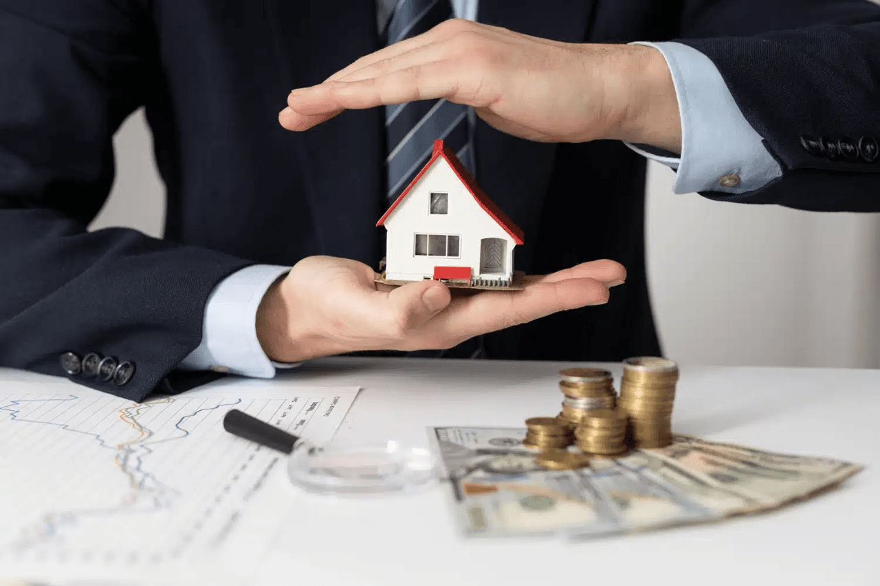 A suited person presents a model house above coins, banknotes, and financial charts.